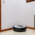 **

A compact robot vacuum cleaner, triangular shaped, effectively cleaning the corner of a small apartment living room in Vietnam. The room has typical Vietnamese decor, like a Phật Di Lặc statue or a conical hat (nón lá) on the wall. Show the robot easily navigating around common obstacles like a power cord and a child's toy.

**
